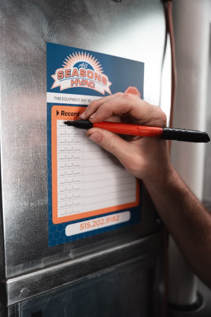 A hand recording maintenance on an All Seasons HVAC service-record label attached to a furnace